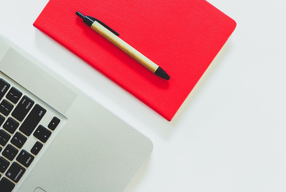 A pen sits on top of a red notebook beside an open laptop.